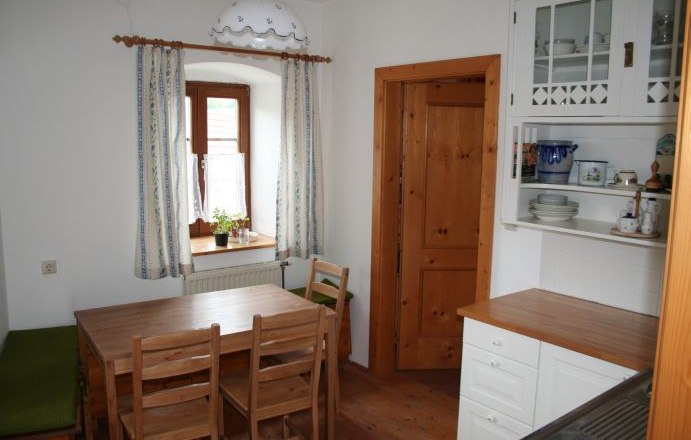 Kleine K&uuml;che mit Holzm&ouml;beln, Esstisch, St&uuml;hlen und einem Fenster mit Vorh&auml;ngen.