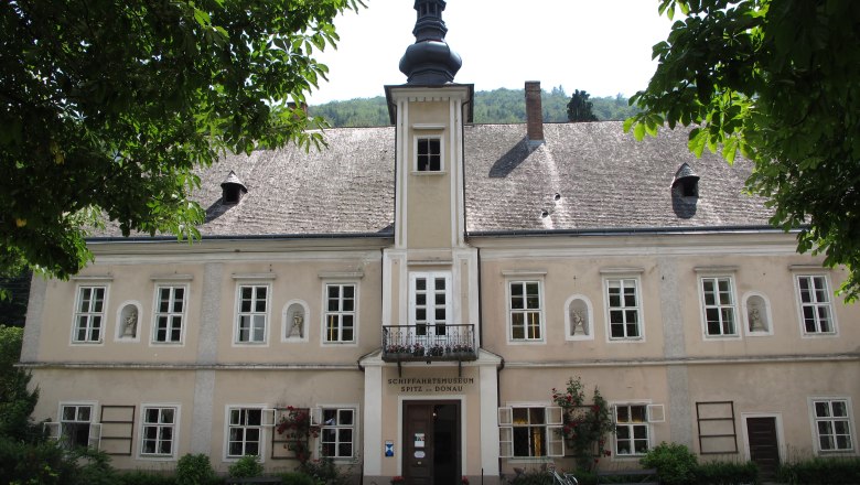 Außenansicht eines historischen Gebäudes mit Schindeldach und Turm, umgeben von Bäumen.