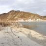 A quiet beach with pebbles, a village and wooded hills in the background.