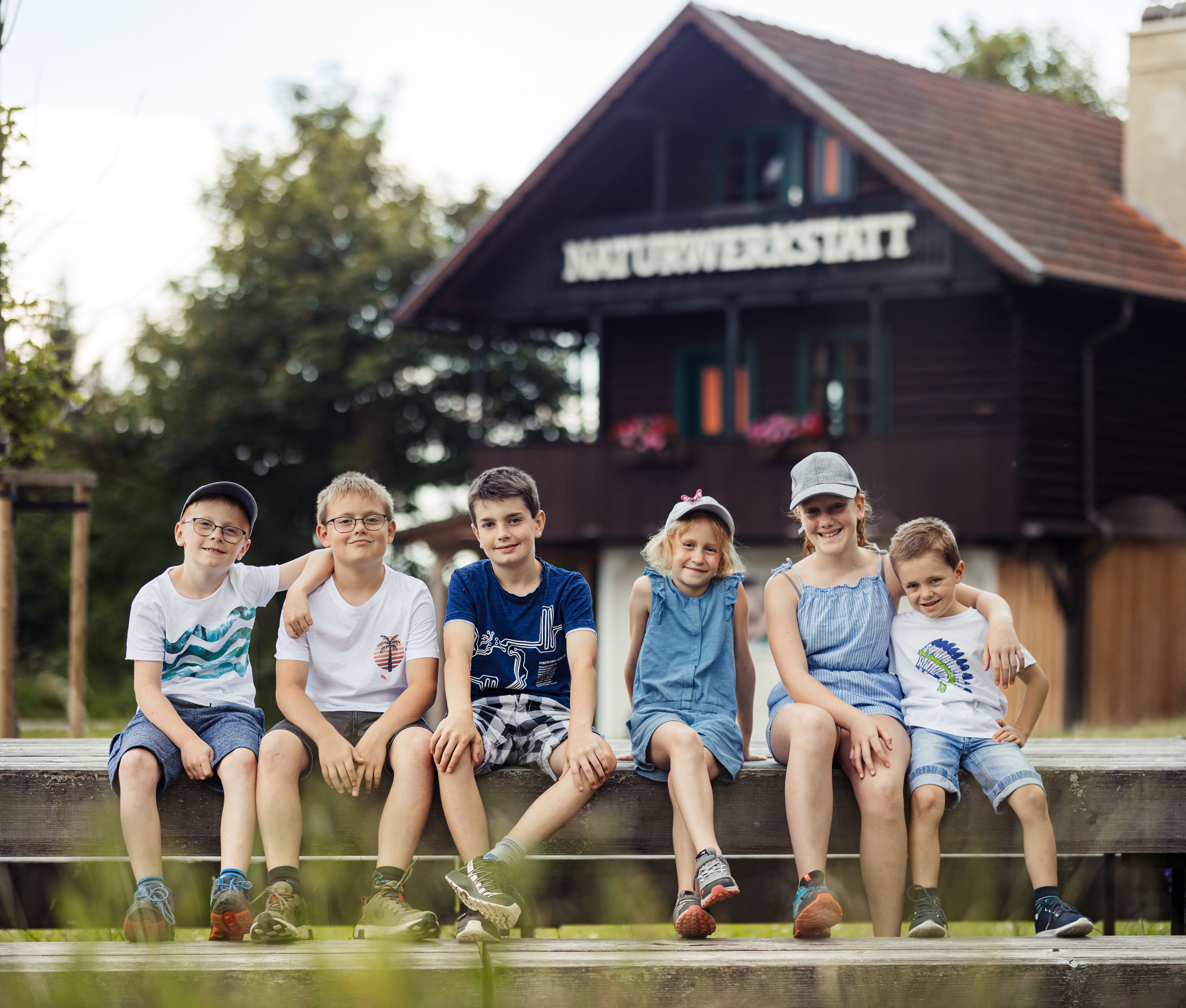 Sechs Kinder sitzen lachend auf einer Holzbank vor einer Holzhütte mit der Aufschrift Naturwekstatt