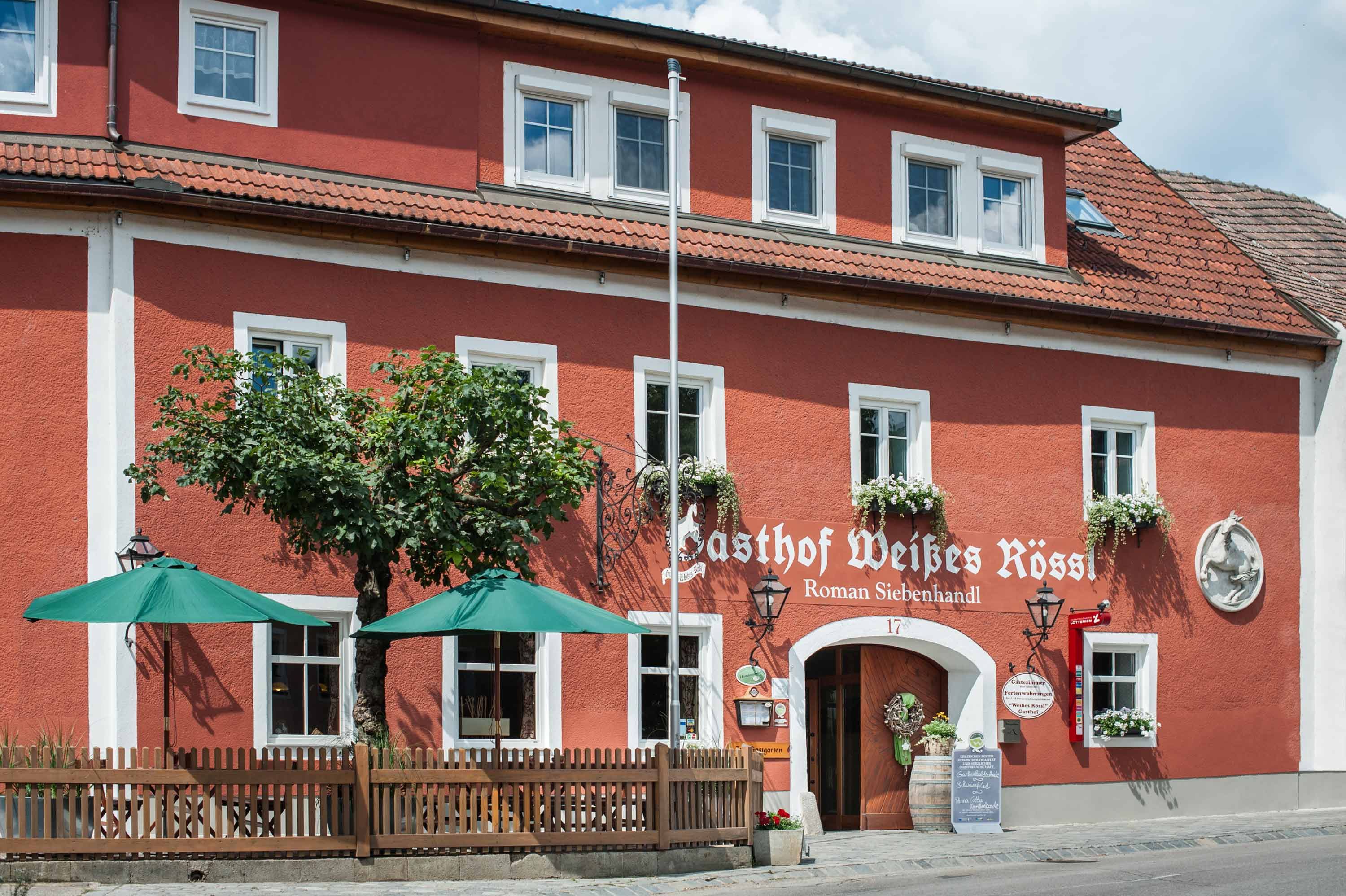 Rotes Gebäude mit weißen Fensterrahmen und grünen Sonnenschirmen vor dem Eingang.