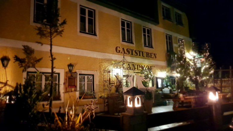 Night view of a yellow building with the inscription 'Gaststuben Landhof', illuminated by lanterns and surrounded by plants.